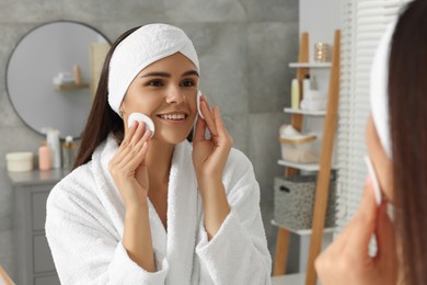 Photo of Young woman cleaning her face with cotton pads near mirror in bathroom