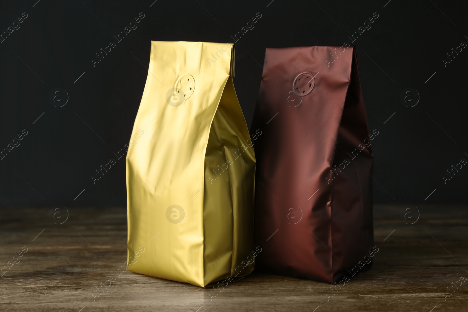 Photo of Different blank foil packages on wooden table against dark background