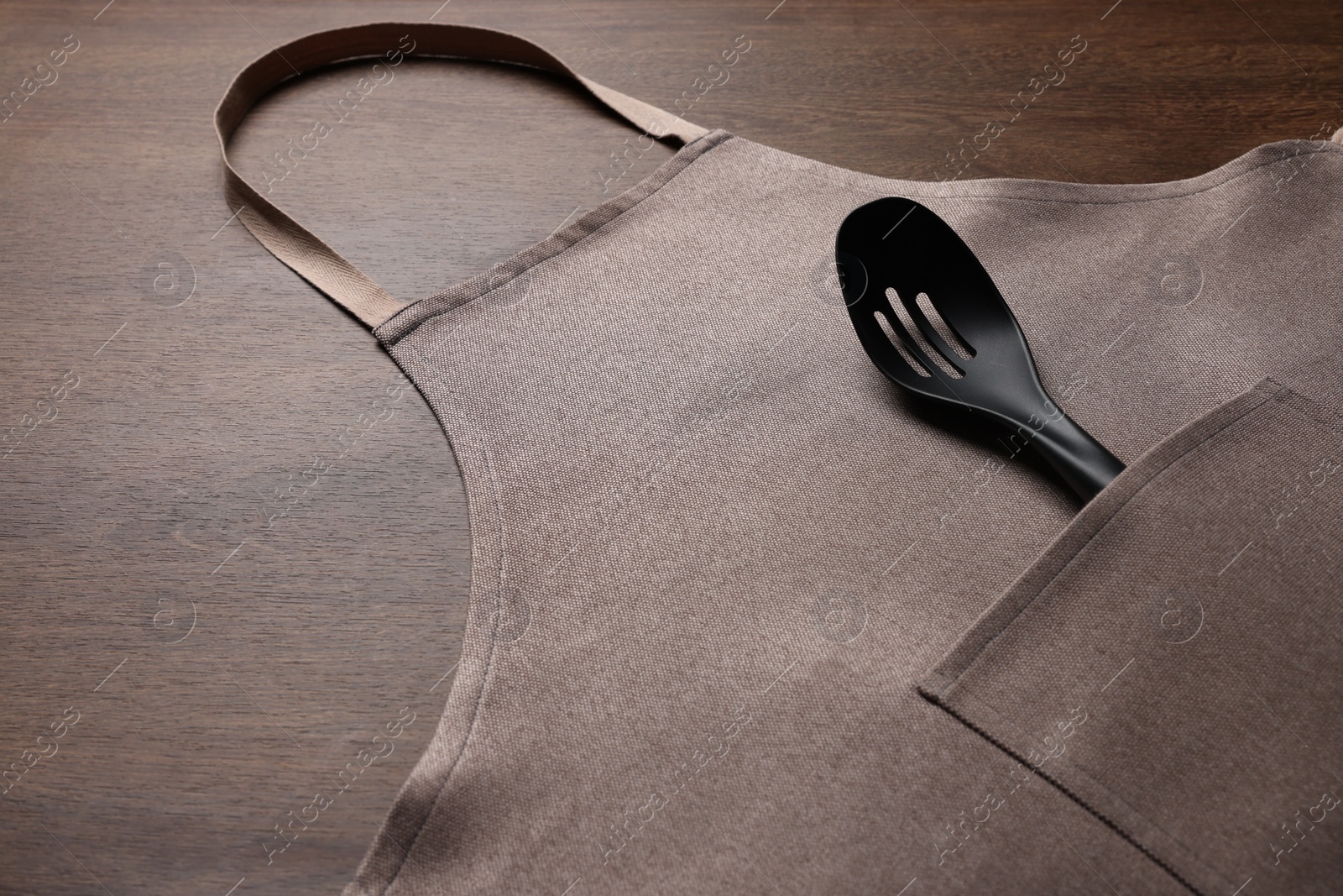 Photo of Stylish brown apron and slotted spoon on wooden table. Space for text