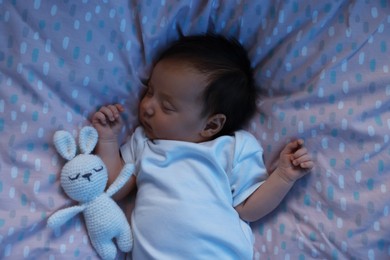 Cute newborn baby sleeping with toy bunny in crib at night, top view