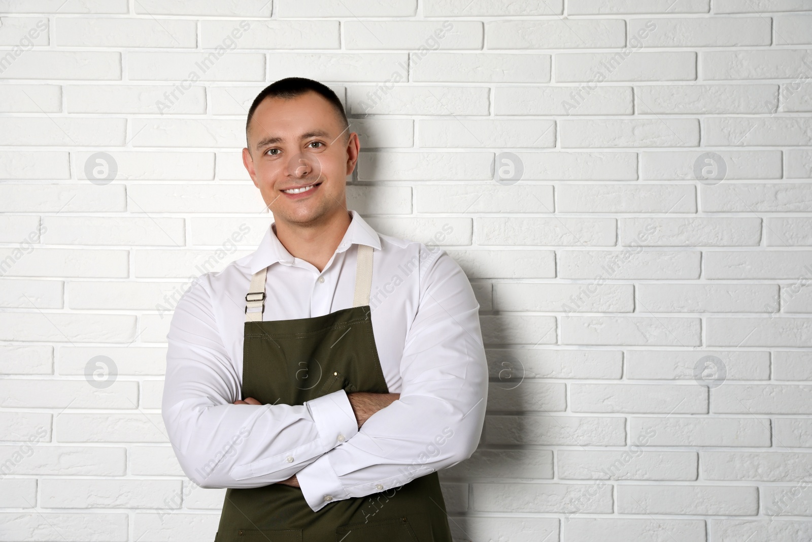 Photo of Portrait of happy young waiter in uniform near white brick wall, space for text