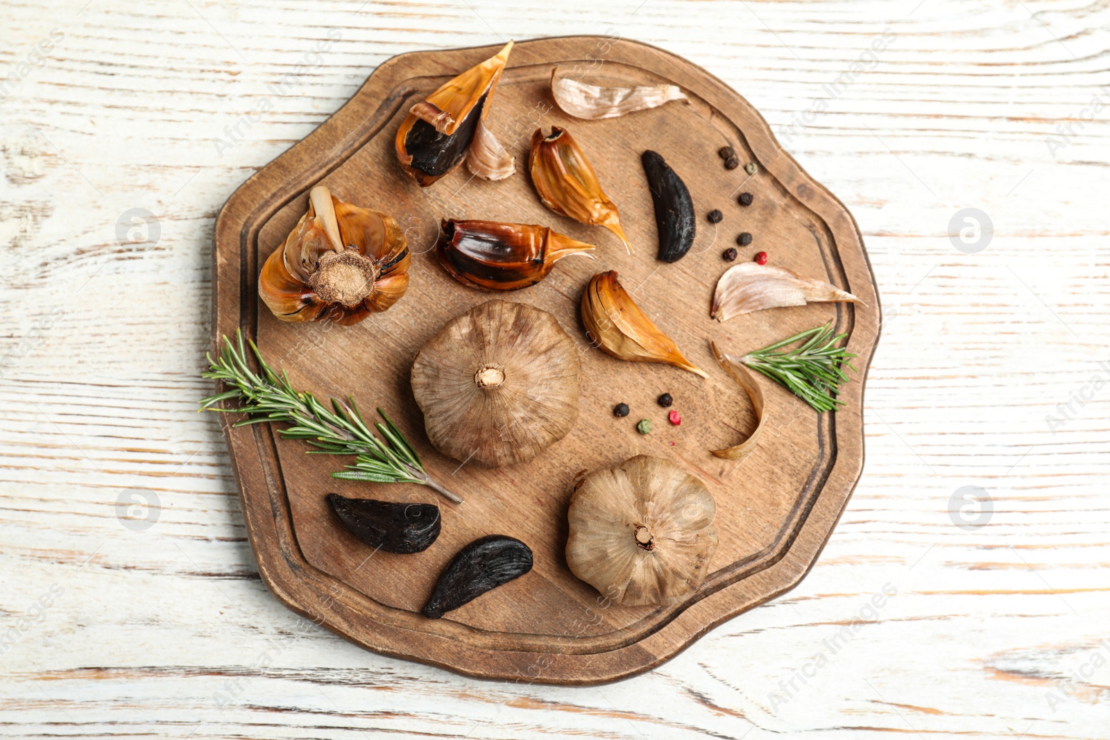 Photo of Board with black garlic and rosemary on white wooden table, top view