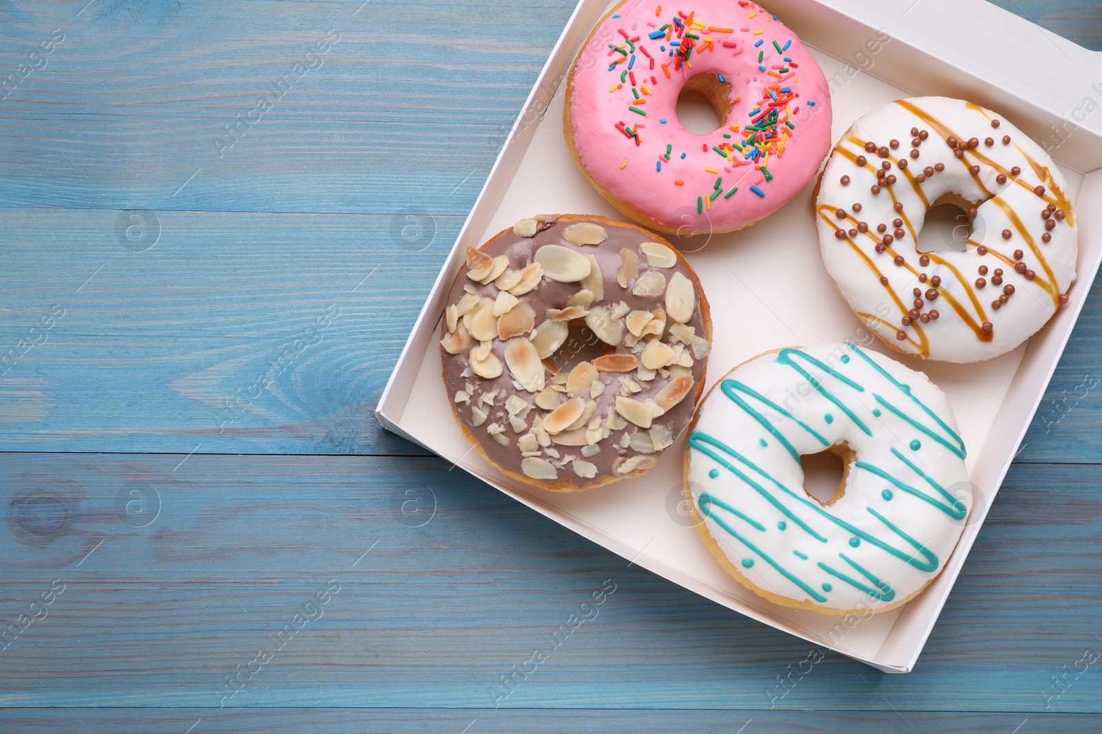 Photo of Box with different tasty glazed donuts on light blue wooden table, top view. Space for text