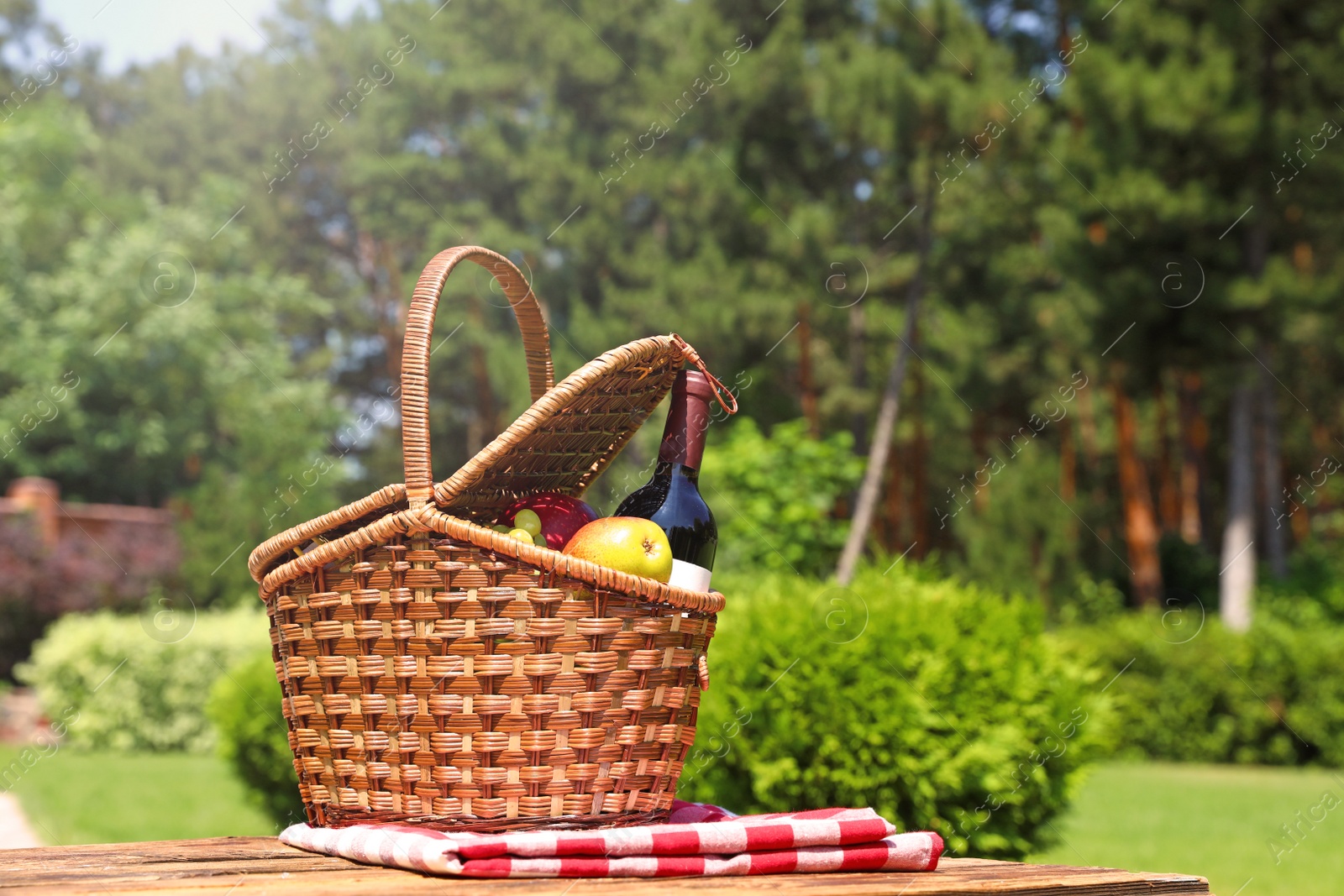 Photo of Picnic basket with fruits, bottle of wine and checkered blanket on wooden table in garden. Space for text
