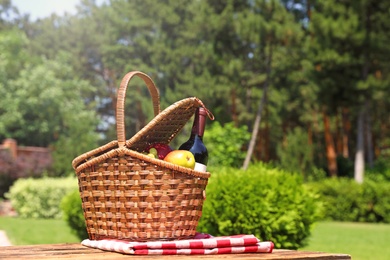 Picnic basket with fruits, bottle of wine and checkered blanket on wooden table in garden. Space for text