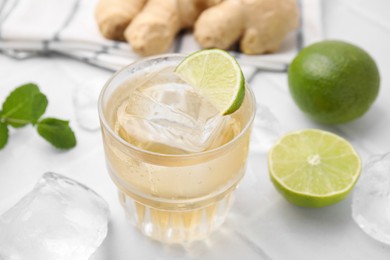 Photo of Glass of tasty ginger ale with ice cube and ingredients on white tiled table, closeup