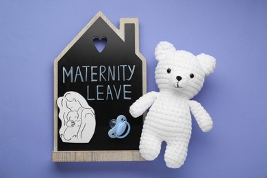 Photo of Maternity leave concept. Wooden house figure, baby pacifier, toy bear and paper cutout on violet background, flat lay
