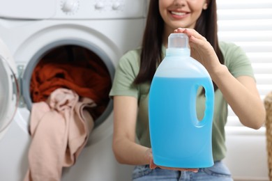 Photo of Woman holding fabric softener near washing machine with dirty clothes, closeup. Space for text