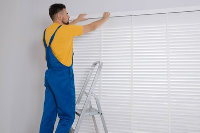 Photo of Worker in uniform installing horizontal window blinds on stepladder indoors