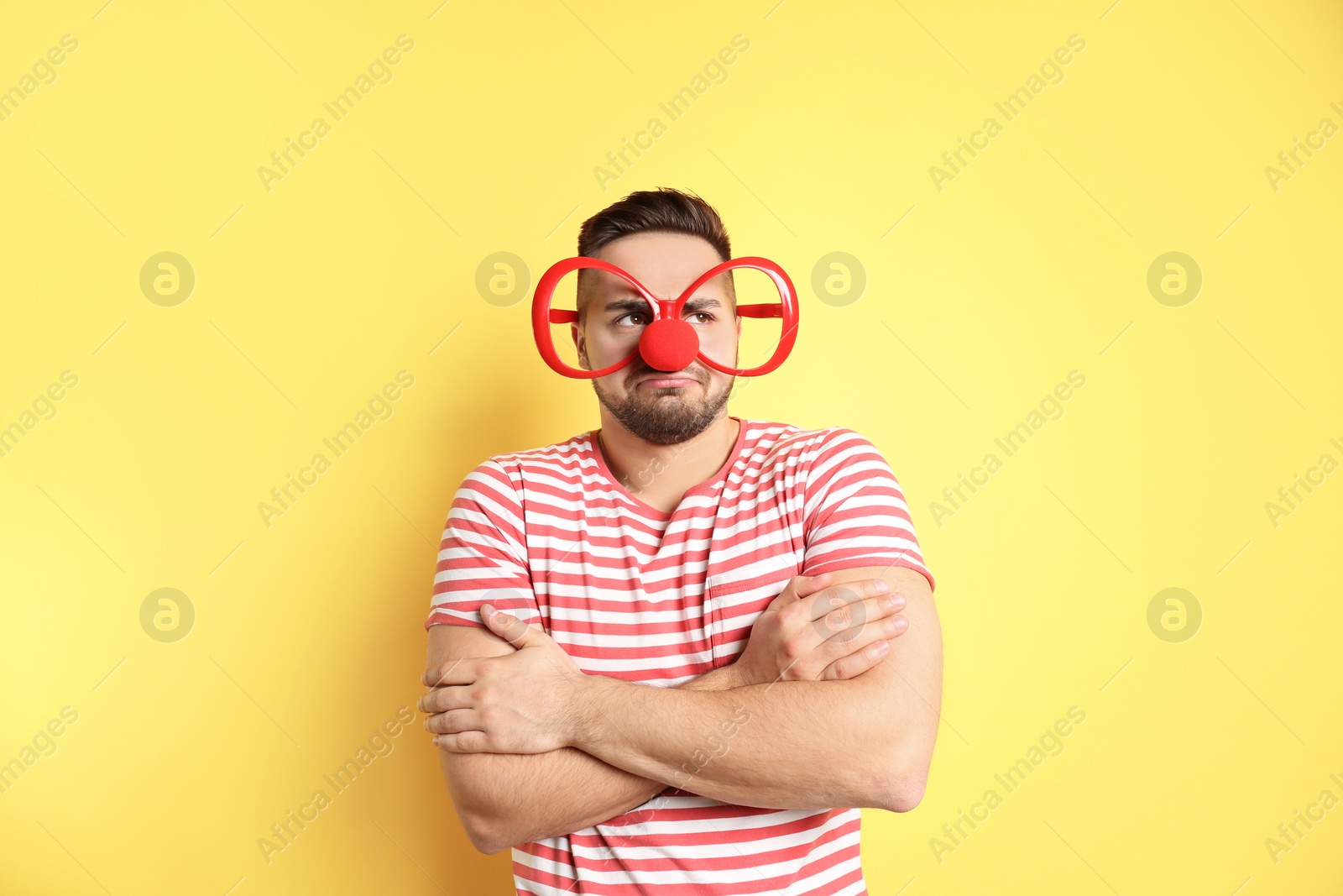 Photo of Emotional young man with party glasses and clown nose on yellow background. April fool's day