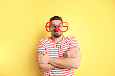 Emotional young man with party glasses and clown nose on yellow background. April fool's day