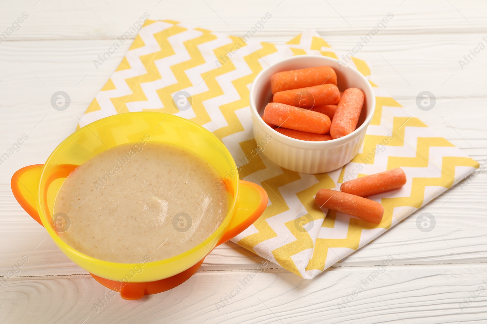 Photo of Baby food. Puree in bowl and small carrots on white wooden table