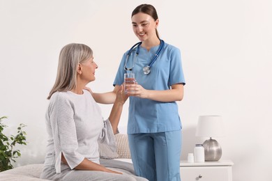 Young healthcare worker giving glass of water to senior woman indoors