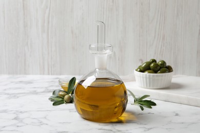 Photo of Glass jug of oil, ripe olives and green leaves on white marble table