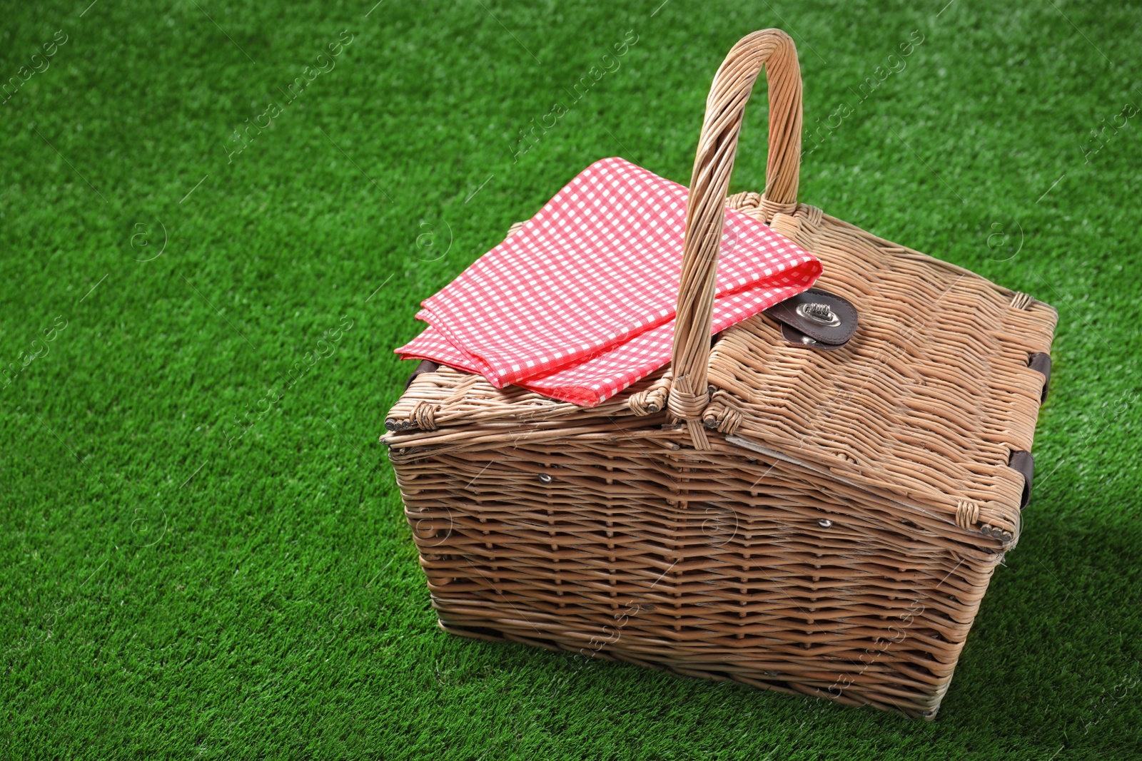 Photo of Checkered tablecloth and closed wicker picnic basket on green grass, space for text