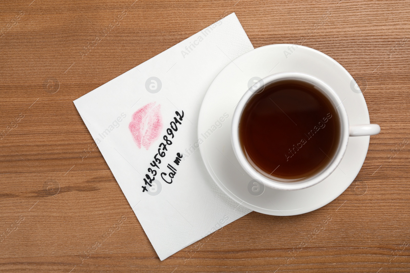 Photo of Paper napkin with phone number and lipstick mark under tea cup on wooden table, top view