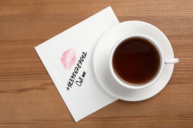 Photo of Paper napkin with phone number and lipstick mark under tea cup on wooden table, top view