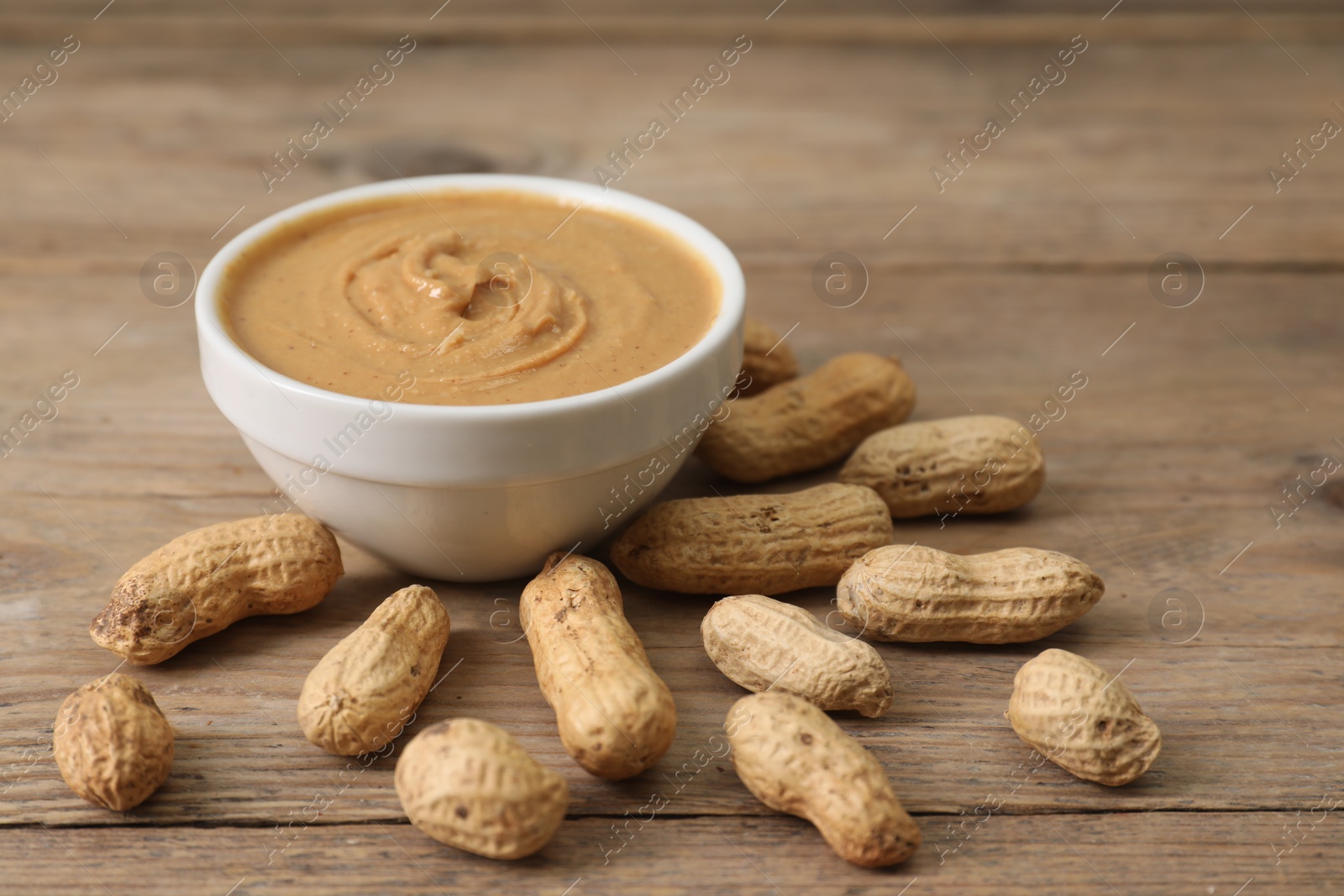 Photo of Delicious nut butter in bowl and peanuts on wooden table, closeup