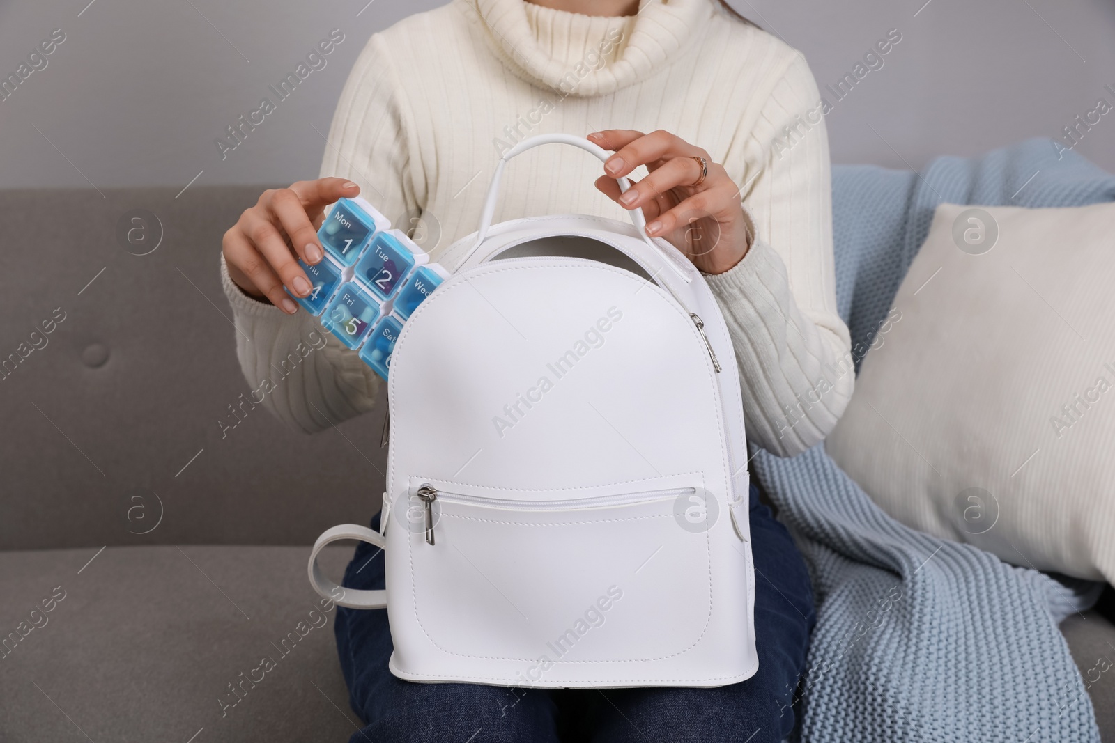 Photo of Woman putting plastic box with pills into white backpack on sofa at home, closeup