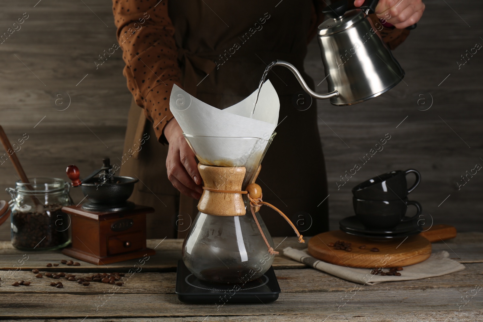 Photo of Woman pouring hot water into glass chemex coffeemaker with paper filter at wooden table, closeup