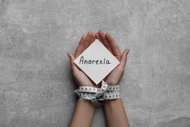 Photo of Woman holding paper note with word Anorexia in hands tied with measuring tape, top view