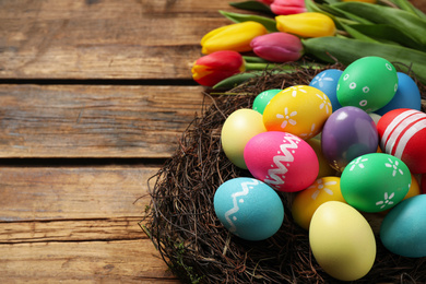 Photo of Colorful Easter eggs in decorative nest and tulips on wooden background, closeup. Space for text
