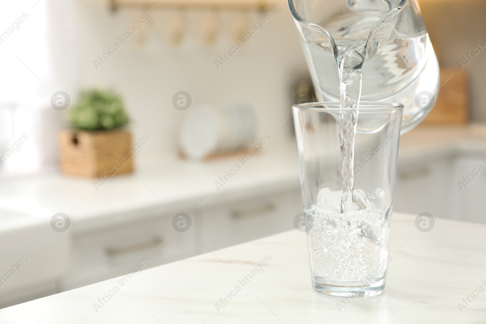 Photo of Pouring water from jug into glass on white table in kitchen. Space for text