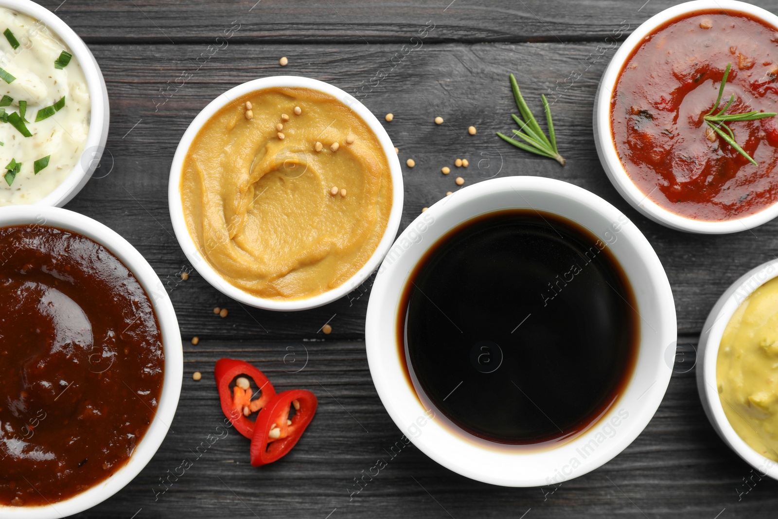 Photo of Different tasty sauces in bowls, parsley, chili pepper and rosemary on black wooden table, flat lay