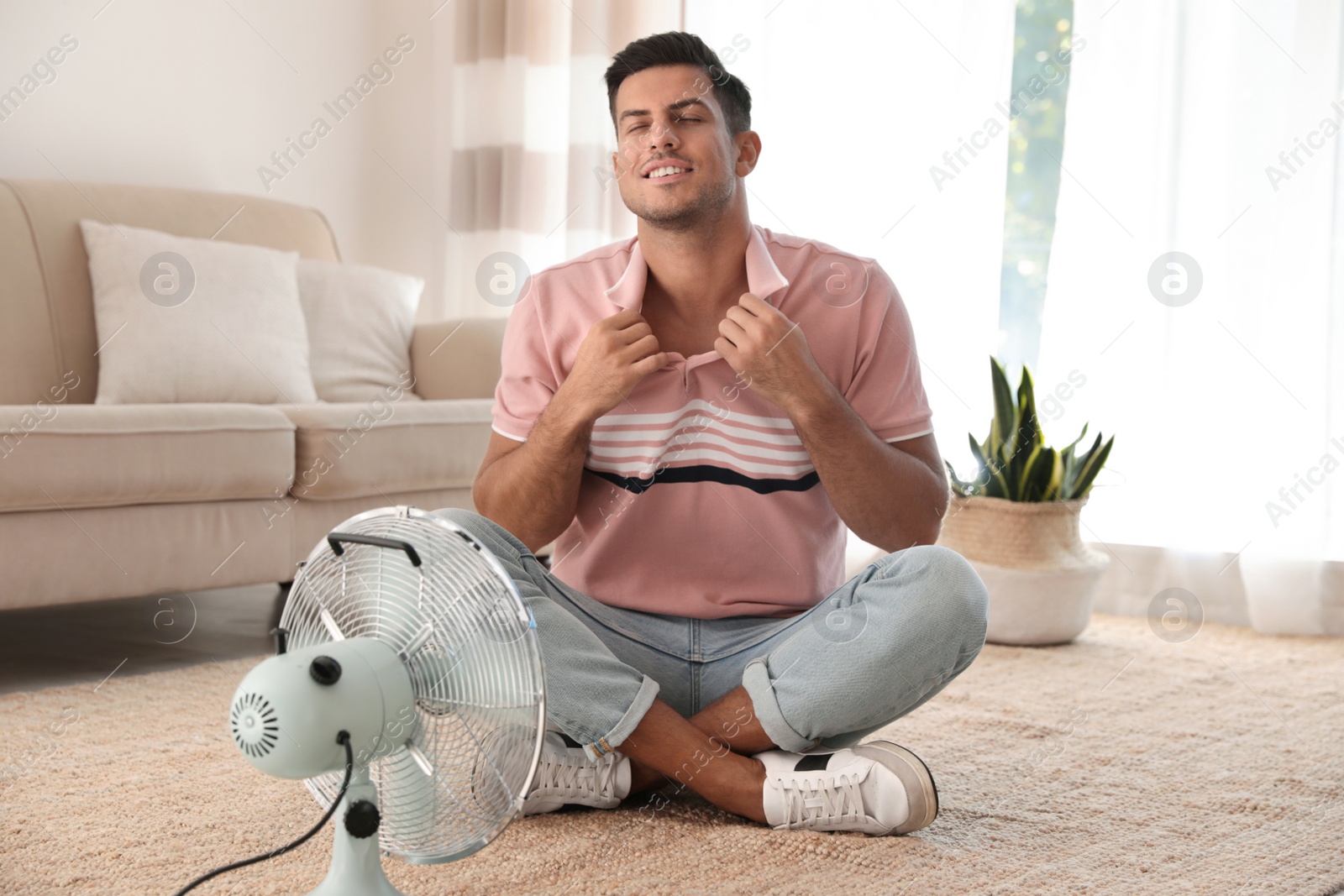 Photo of Man enjoying air flow from fan on floor in living room. Summer heat