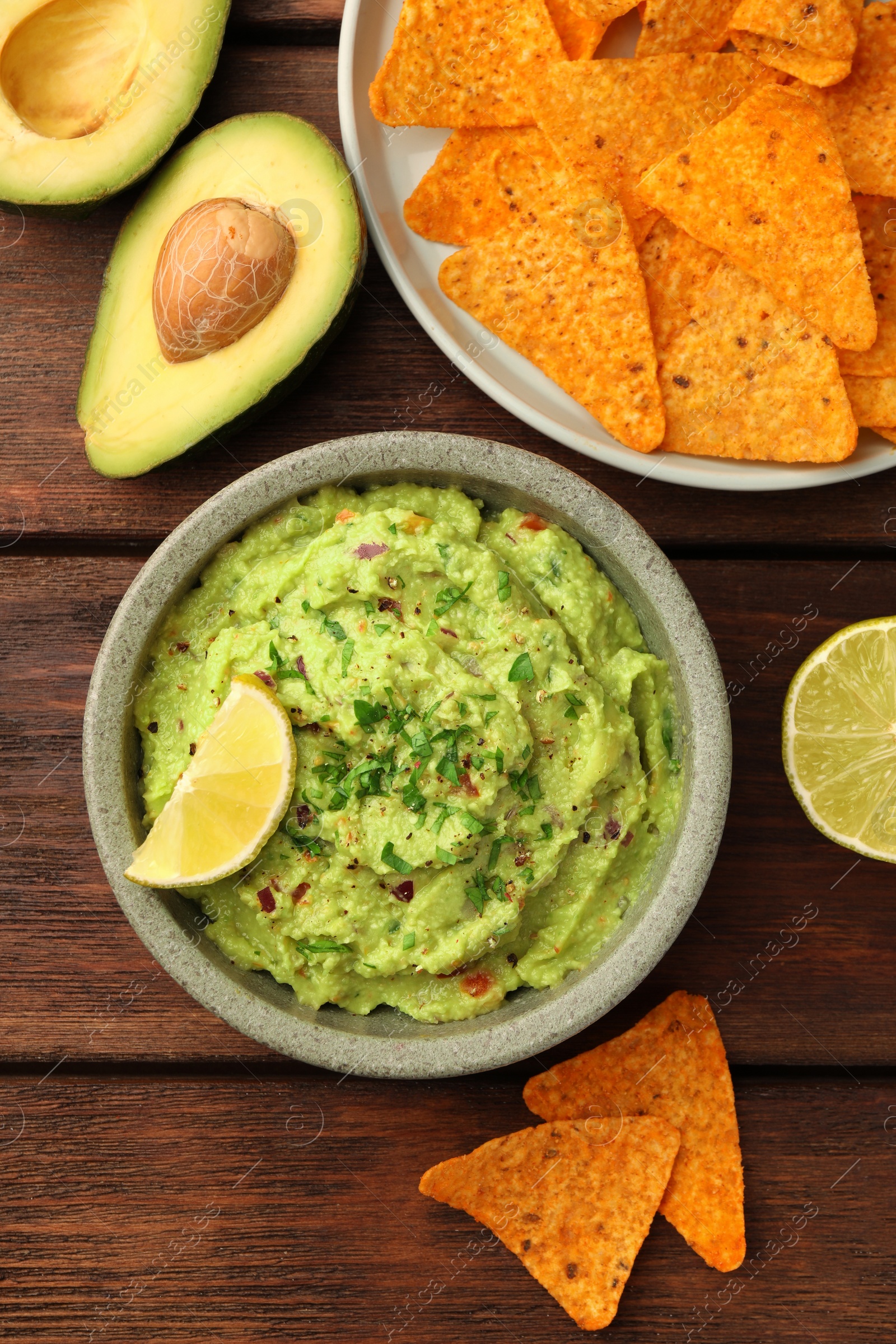 Photo of Bowl of delicious guacamole, lime and nachos chips on wooden table, flat lay