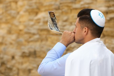 Photo of Jewish man in kippah and tallit blowing shofar outdoors. Rosh Hashanah celebration