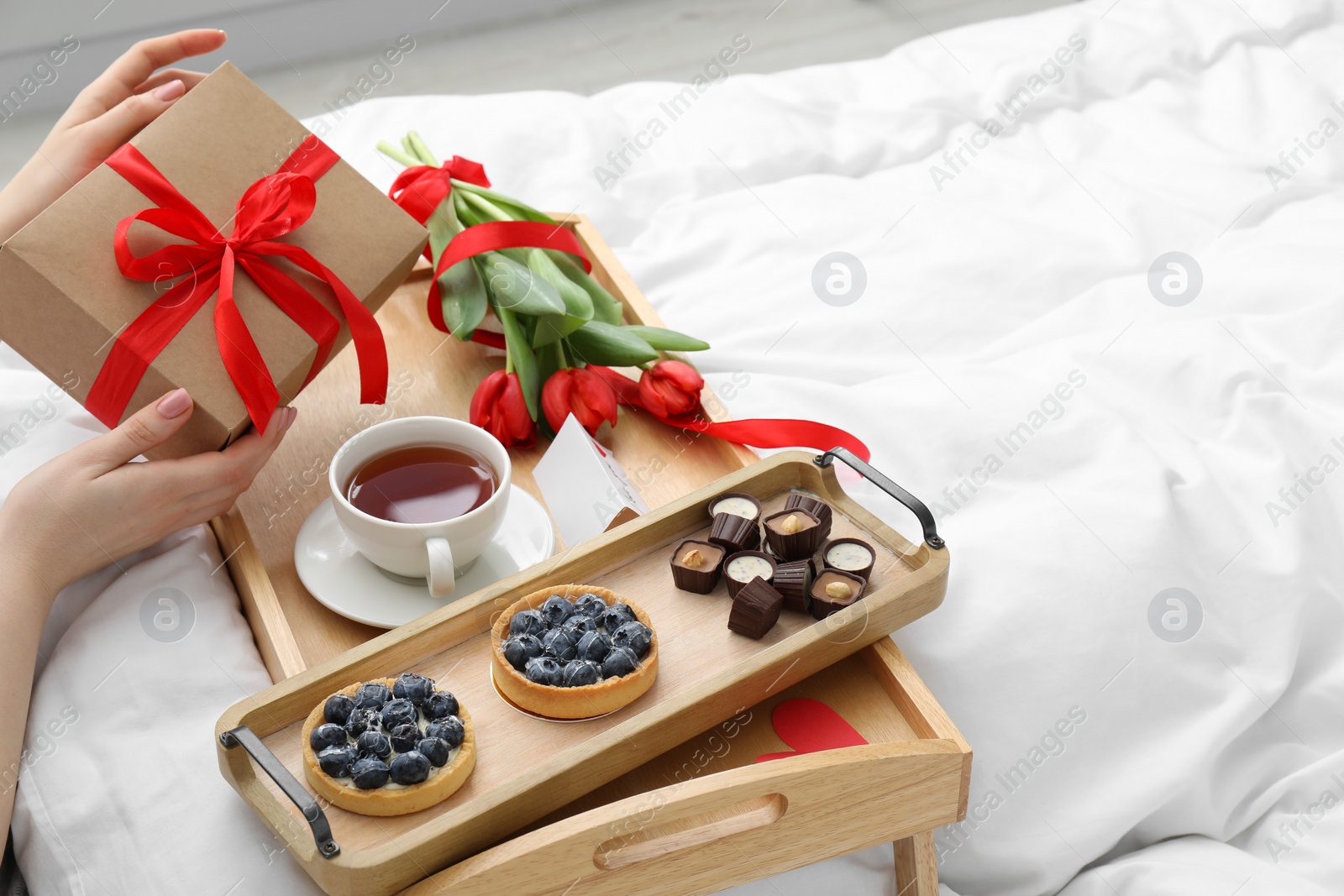 Photo of Tasty breakfast served in bed. Woman with gift box, desserts, tea and flowers at home, closeup. Space for text