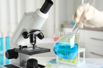 Medical assistant holding glassware with liquid near modern microscope in laboratory, closeup