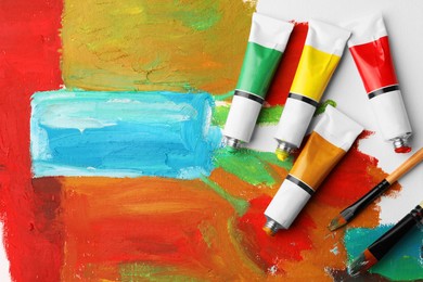 Photo of Tubes of colorful oil paints and brushes on canvas with abstract painting, flat lay