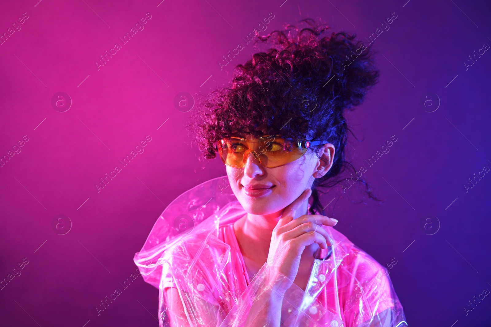 Photo of Beautiful young woman in transparent coat and sunglasses posing on color background in neon lights