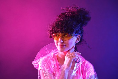 Photo of Beautiful young woman in transparent coat and sunglasses posing on color background in neon lights