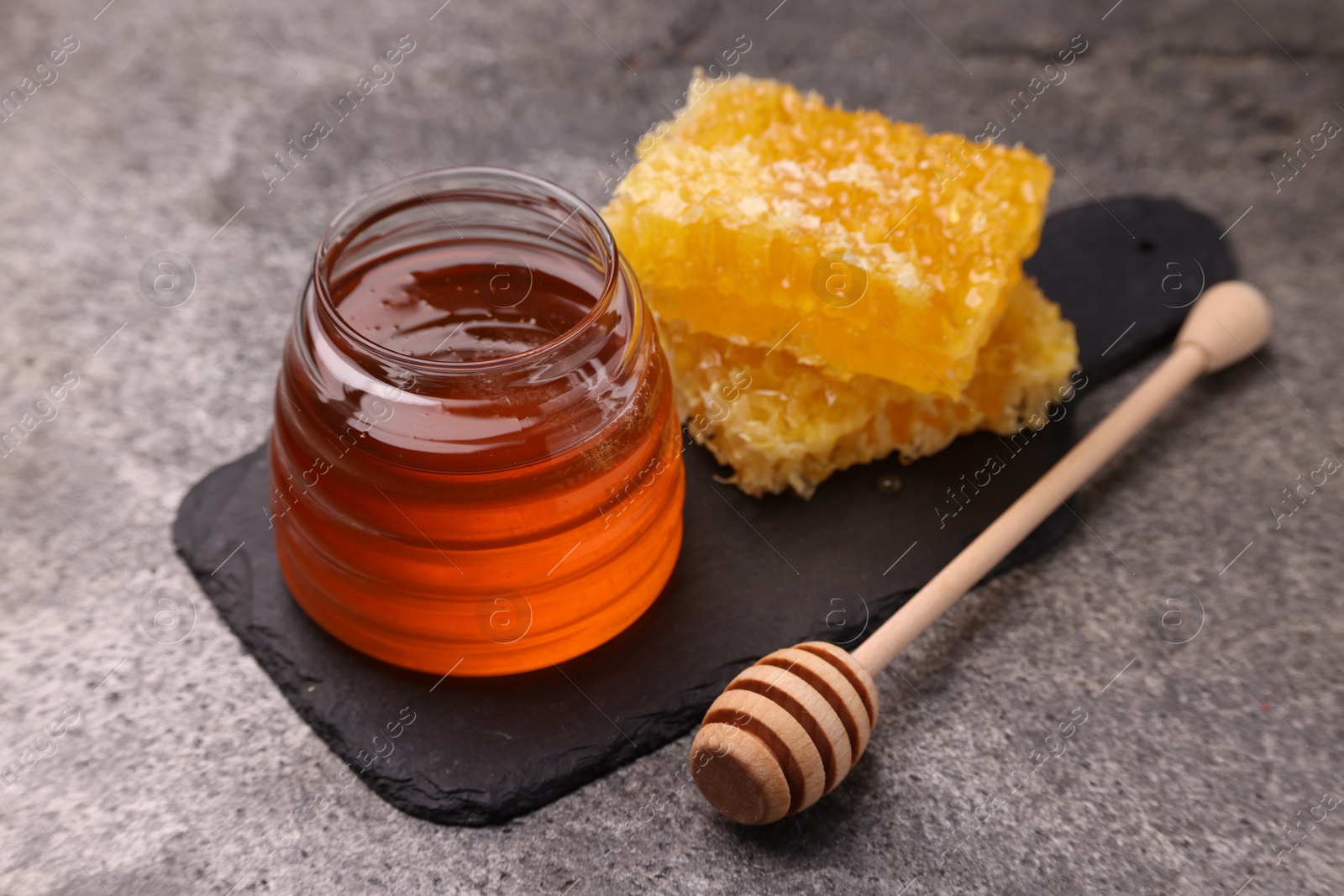 Photo of Sweet golden honey in jar, dipper and pieces of honeycomb on grey textured table