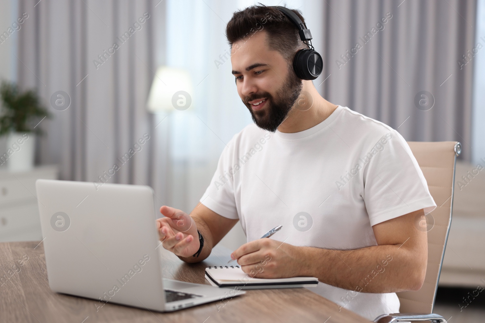 Photo of Young man in headphones using video chat during webinar at table in room