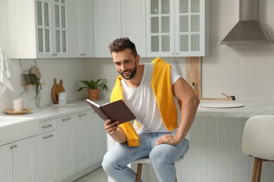 Photo of Handsome man reading book on stool in kitchen
