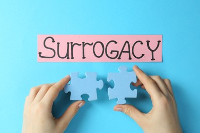 Woman holding puzzle pieces near sheet of paper with word Surrogacy on light blue background, top view