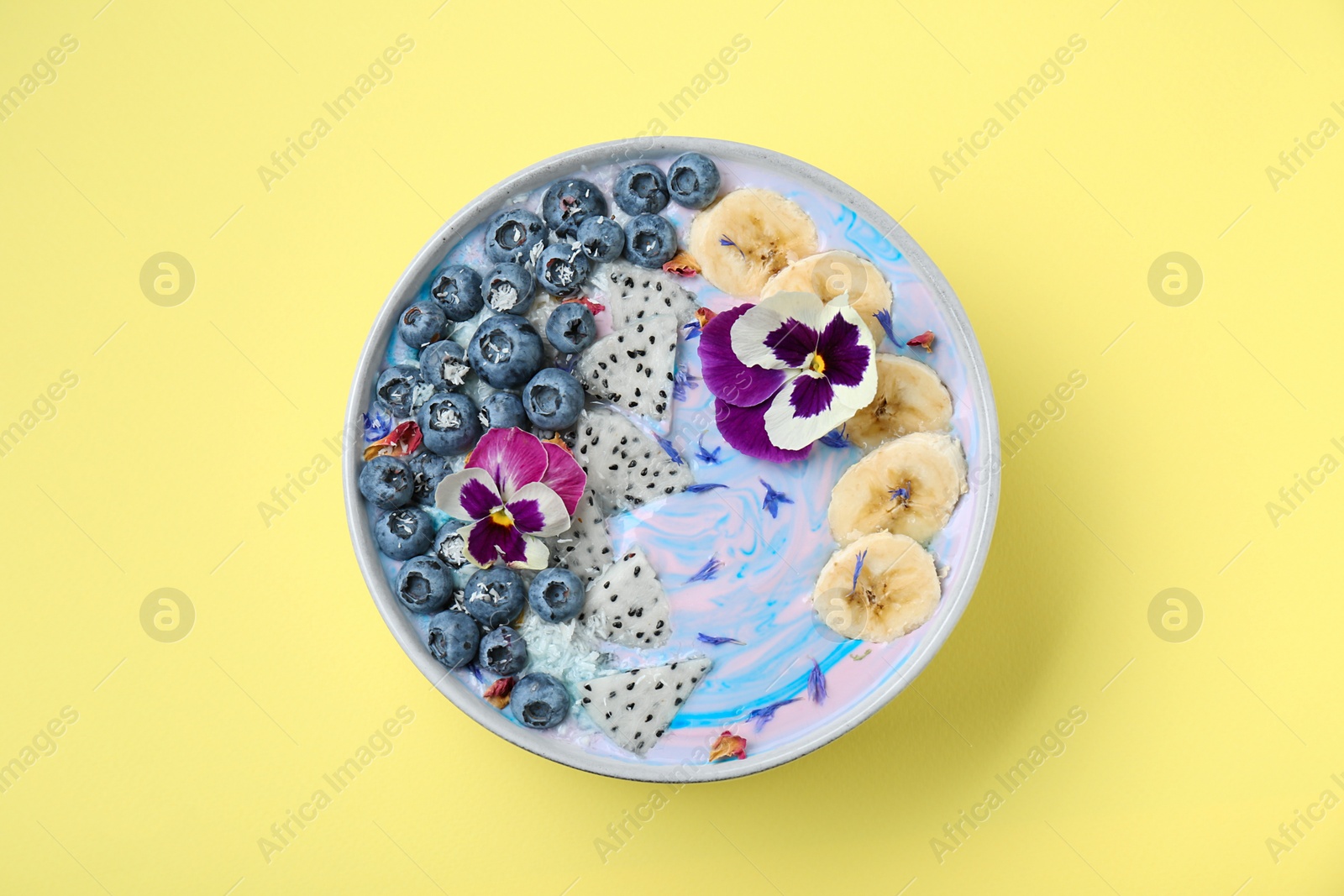 Photo of Delicious smoothie bowl with fresh fruits, blueberries and flowers on yellow background, top view