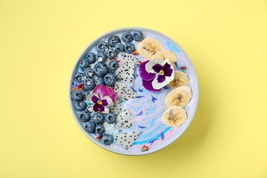 Photo of Delicious smoothie bowl with fresh fruits, blueberries and flowers on yellow background, top view