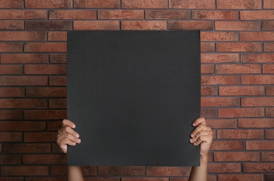 Man holding black blank poster near red brick wall, closeup. Mockup for design