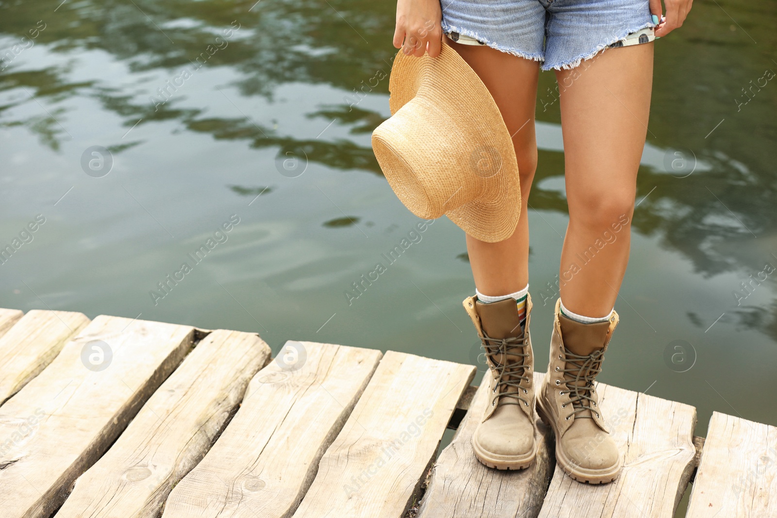 Photo of Young woman with hat on wooden pier near lake, focus on legs. Camping season