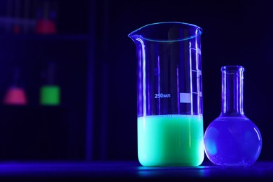 Laboratory glassware with luminous liquids on table against dark background, space for text