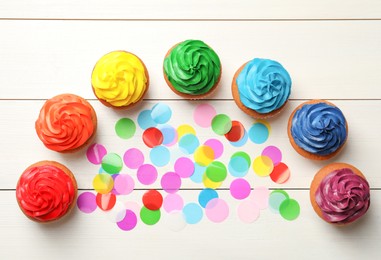 Photo of Many delicious colorful cupcakes and confetti on white wooden table, flat lay