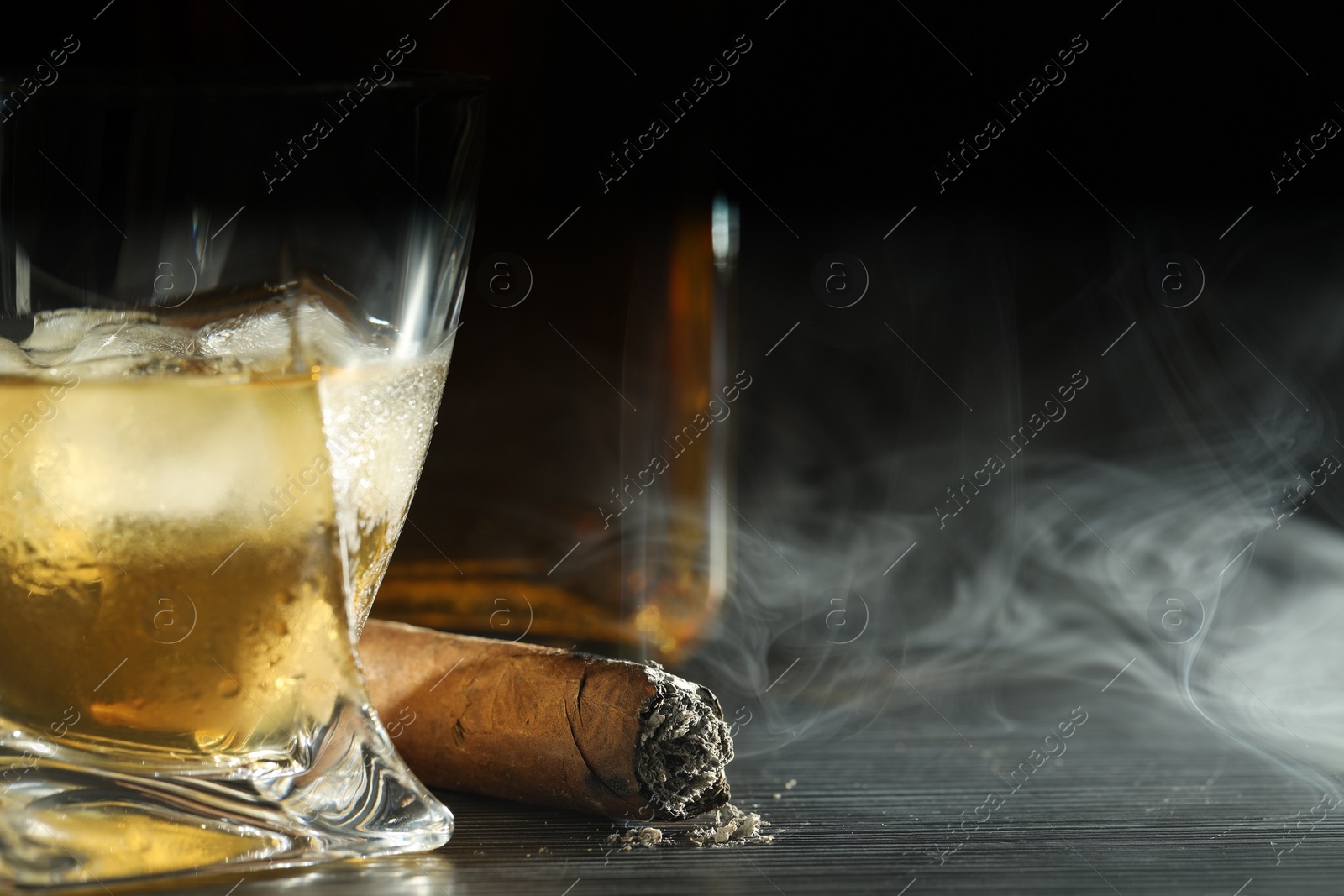 Photo of Glass of whiskey with ice cubes and smoldering cigar on black wooden table, closeup. Space for text