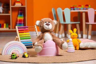 Photo of Teddy bear on pink baby potty and many other toys in room. Toilet training