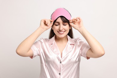 Happy woman in pyjama and sleep mask on light grey background