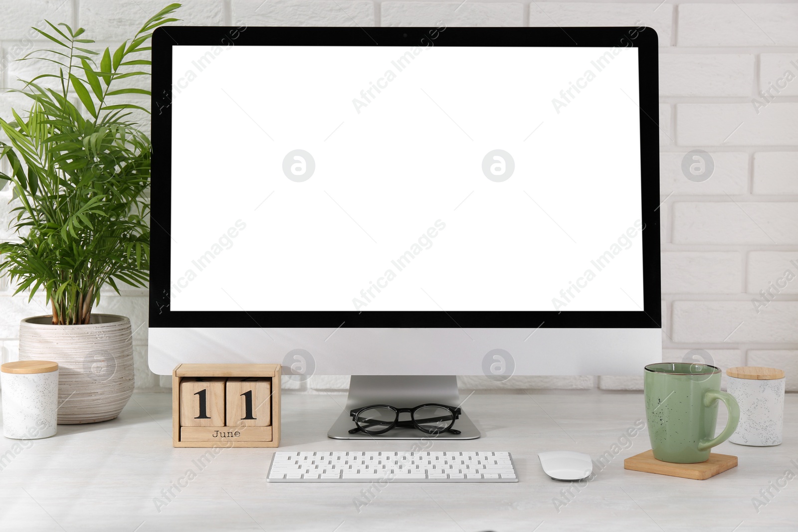 Photo of Office workplace with computer, glasses, cup, wooden block calendar and houseplant on light table near white brick wall